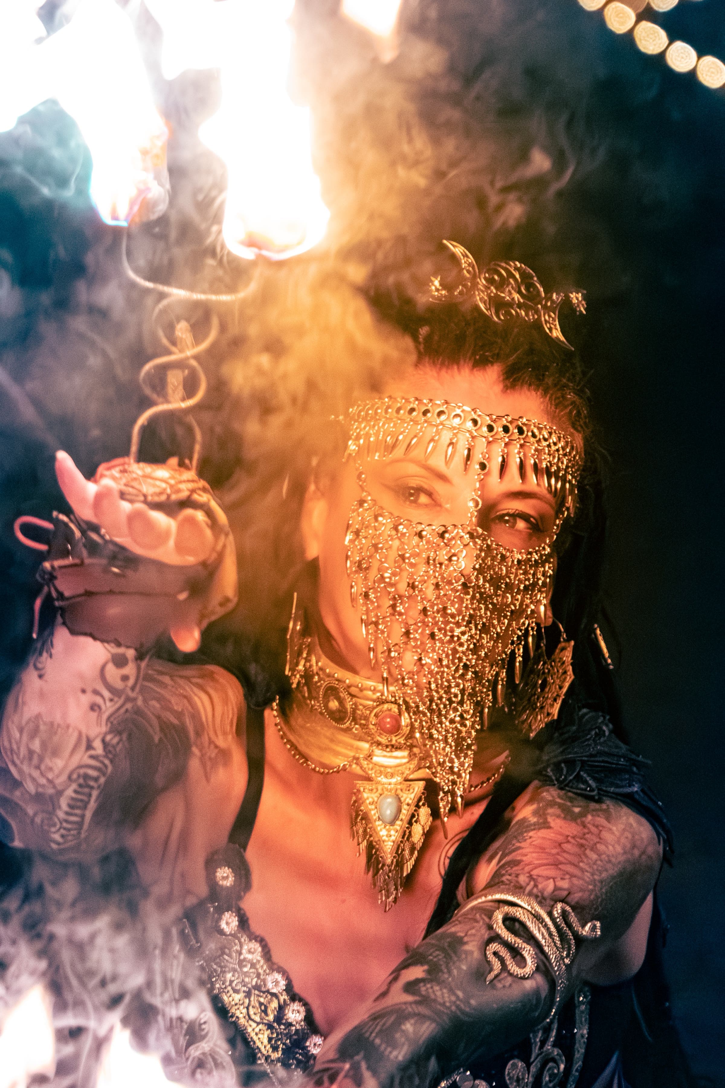 A fire performer in a gold chainmail veil and crescent headpiece, holding a flaming prop amid smoke and warm firelight.