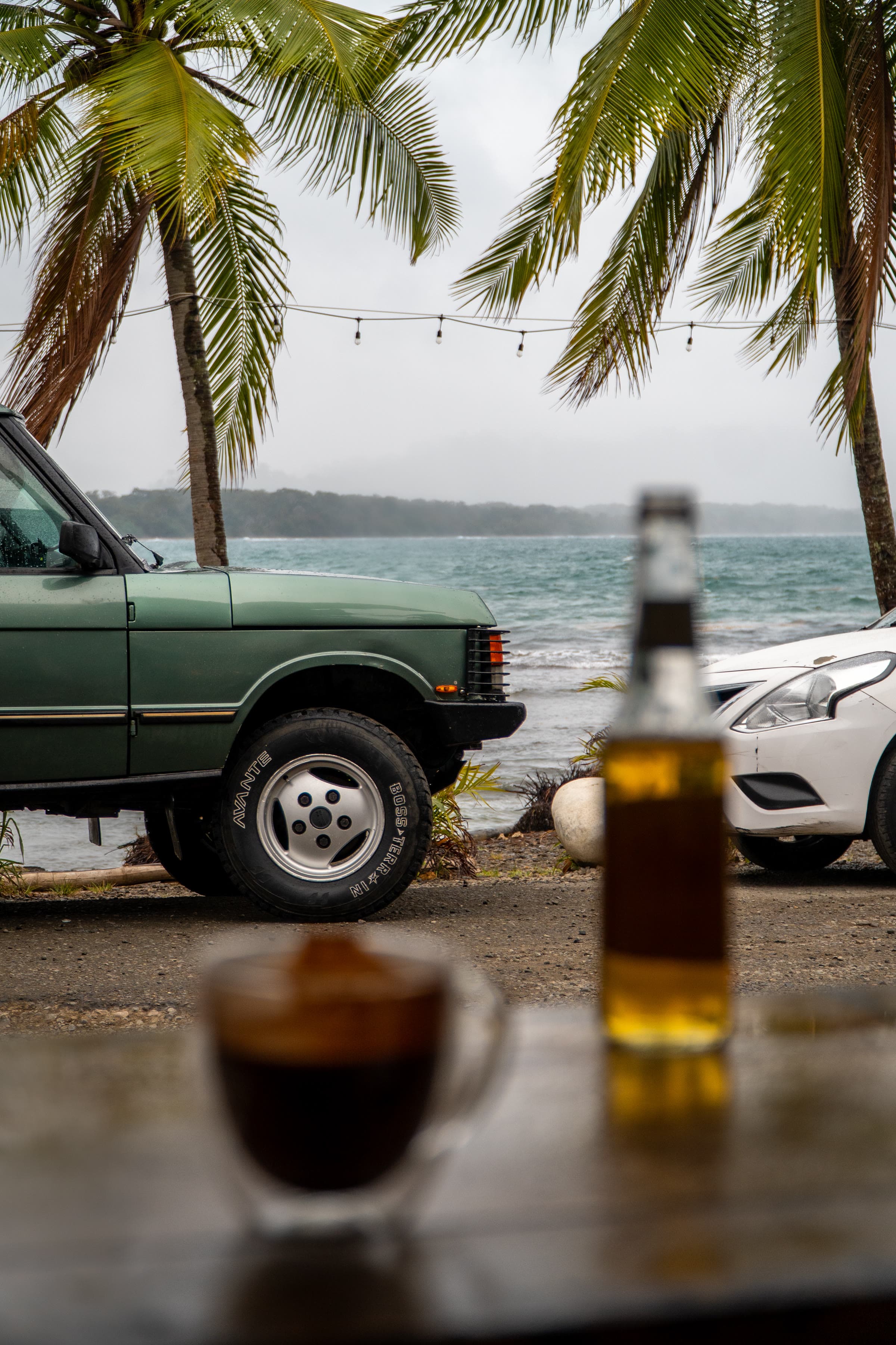 Sage-green Range Rover on a gravel shore beside turquoise water, under palm fronds and festoon lights, with coffee and a bottle on a wooden ledge in front.